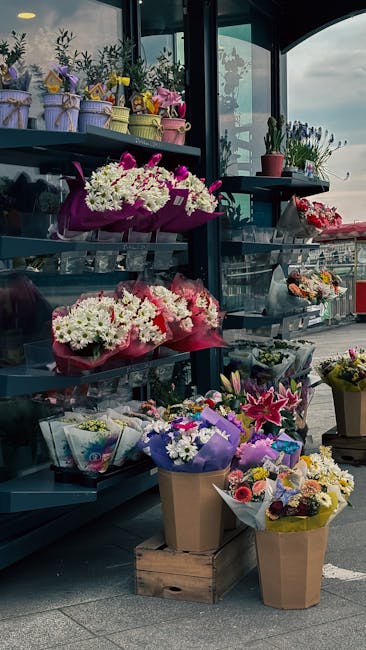 A display of various floral arrangements on metal shelves inside a flower shop, featuring bouquets wrapped in pink, purple, and brown paper with a mix of white, pink, yellow, and purple flowers. The a