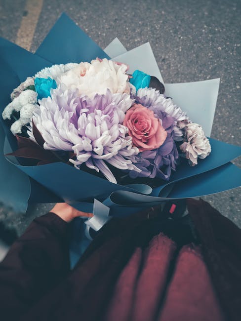 A floral bouquet featuring a mix of large, white and light purple chrysanthemums, soft pink roses, and smaller blue and white accent flowers, wrapped in elegant blue and white paper with a glossy fini