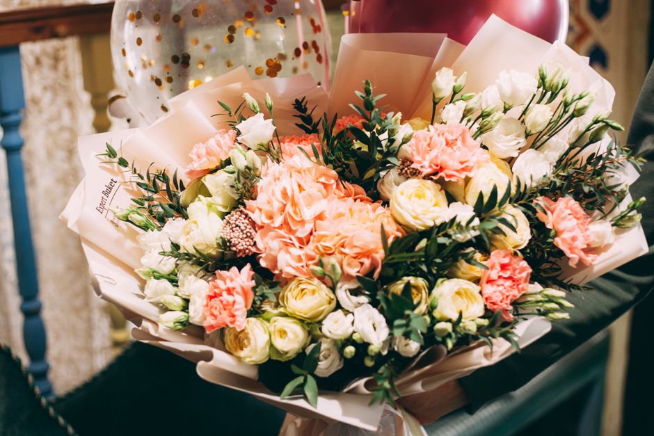 A vibrant floral bouquet featuring a mix of peach and white roses, delicate white lisianthus, and blush pink carnations, arranged with lush green foliage. The flowers are wrapped in elegant pale pink