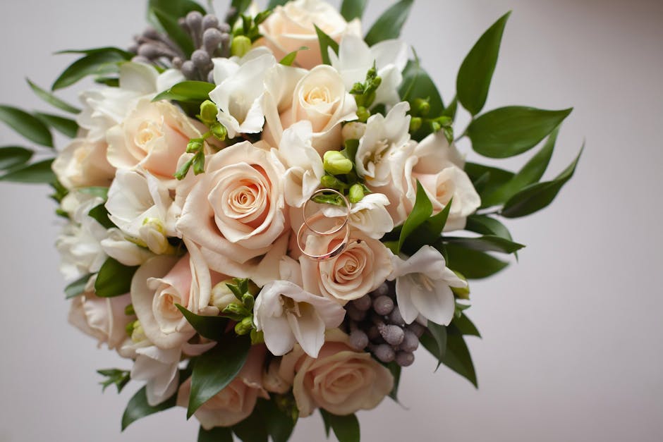A close-up image of a romantic floral bouquet featuring soft blush pink roses, white freesias, and small white blossoms, accented with green leaves and sprigs of greyish lavender, arranged in a round,