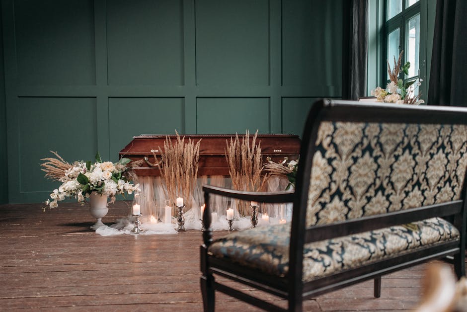 A floral arrangement placed on a wooden table in an indoor setting, featuring tall dried pampas grass and white flowers arranged in a vase, surrounded by small lit candles in silver holders and soft w