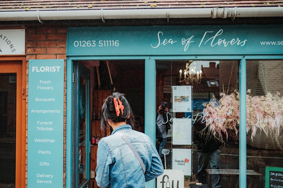 A woman with dark hair pinned with a bright red clip stands outside the storefront of 'Sea of Flowers,' a floral shop with a teal sign and window frame. She is wearing a light denim jacket and carries