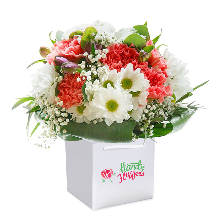 Set elegantly against a backdrop of pure white, a richly textured bouquet sits poised in a squared white gift bag, its composition and colour harmonies capturing the vibrant energy and calm beauty unique to Kidbrooke. The main attraction-sumptuous coral and peach carnations-unfolds in soft, overlapping layers, their ruffled petals lending a fullness as inviting as a walk through Kidbrooke Green Park on a fresh spring day. Explosions of bright white daisy-like chrysanthemums, each center a punch of juicy chartreuse, frame the arrangements with light and optimism. Swirling amidst the blooms, tiny clouds of baby's breath bring an ethereal whimsy, their airy touch recalling the gentle breezes over Thomas Tallis School fields. A few tightly closed, mauve-tinged buds insert subtle tension and visual intrigue along the bouquet's left edge, peeking coyly out and suggesting a story yet to unfold. Ringing the flowers, lavish dark green foliage contributes polish and natural support, creating a sturdy, elegant base. The white satin ribbon handles arch gracefully above, practical for deliveries to homes in Kidbrooke Village or quick pickups along Shooters Hill Road. The bouquet's lush, round shape-bursting with freshness and care-embodies the value placed on thoughtfulness and celebration throughout Kidbrooke, making it an ideal companion for birthdays, special milestones, or everyday acts of kindness.