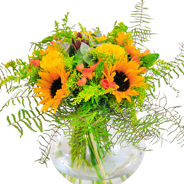 A glance at this bouquet brings to mind high summer walks through Kidbrooke's abundant parks, where golden sunflowers and richly layered yellow roses bloom in harmonious profusion. With their large, round faces and burnt umber centers, the sunflowers convey a sense of happiness and optimism, while spiraled garden roses flaunt a buttery glow reminiscent of the sunset on Kidbrooke Green. Among these sunny blossoms, vibrant clusters of crimson hypericum berries catch the eye, adding depth and a spirited contrast against the warm palette. Lush ferns and gently arching sprigs of goldenrod infuse the arrangement with graceful lines and varied texture, much like the undergrowth found along Shooters Hill Road. Blue-green eucalyptus leaves, with their distinctive silvery cast, filter softly throughout, bringing a fresh, herbal fragrance. The rounded glass vase reveals water-lustred stems, completing the balanced, abundant composition. This bouquet, lovingly arranged by local Kidbrooke florists, is an embodiment of cheerful celebration-perfect for birthdays, graduations, or simply sharing a bright moment with family and friends across SE3.