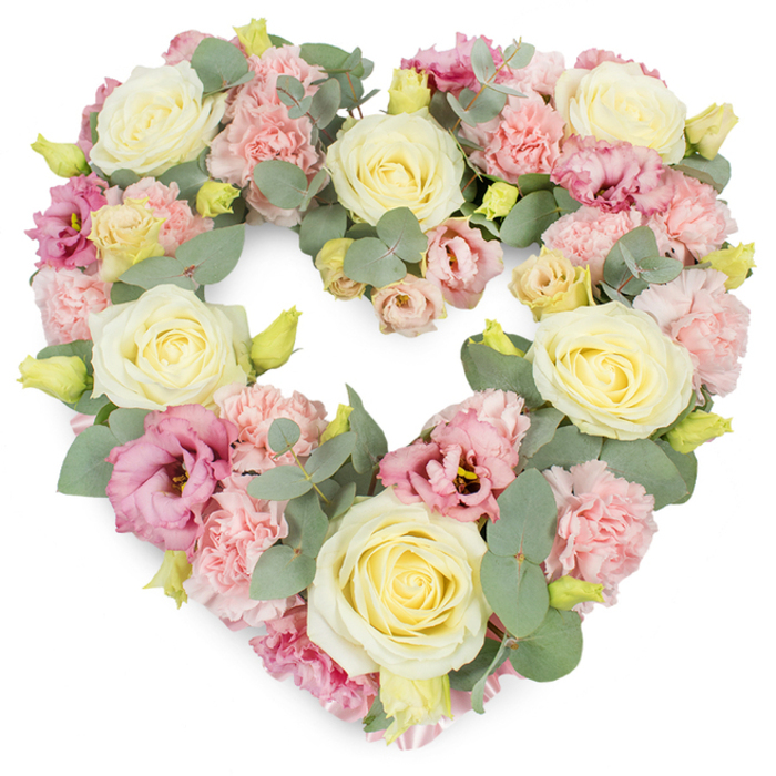 A beautifully balanced, heart-shaped wreath dominates the image, shown against a plain white background that accentuates its soft pastel tones. This is a classic funeral or memorial tribute, with every part of the heart carefully filled, as though no space has been left for emptiness. Cream and pale yellow roses are the stars of the arrangement, positioned evenly across the heart. Their velvety petals spiral out from tight centres, creating rounded blooms that feel calm and reassuring, like the steady presence of someone much loved. Between them, clusters of light pink carnations bring a contrasting, ruffled texture. Their frilly heads sit snugly side by side, giving a sense of volume and gentle movement, as if the flowers are quietly breathing. Among these pastels, deeper pink and mauve lisianthus blossoms provide subtle shadows of colour. Some are fully open, revealing soft folds and sometimes darker centres; others are still in bud or gently cupped, adding depth and visual interest. Cool, silvery eucalyptus leaves weave throughout, their rounded shapes and muted blue-green colour calming the eye and subtly defining the heart's curves. Tiny yellow-green buds punctuate the wreath, adding light touches of vibrancy, like small sparks of memory. The overall mood is peaceful and reflective, perfectly suited to a funeral, cremation ceremony, or celebration of life around Kidbrooke, Eltham, or Blackheath. It feels like the work of a skilled local florist, attuned to the emotions of grieving families, offering a heart-shaped symbol of sympathy, gratitude, and enduring affection that words might struggle to convey alone.