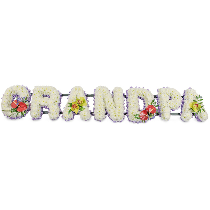 A long line of floral lettering in this photograph spells the word "GRANDPA," forming a striking yet tender focal point suitable for a funeral or graveside tribute. The letters are three-dimensional and robust, but every visible surface is softened by an intricate covering of small white chrysanthemums, each bloom carefully placed so that the petals form a dense, cushiony texture. The white colour reads as clean and bright against the plain background, suggesting peace, reverence, and the simplicity of sincere love. Neatly finishing each letter, a fine edging of pale lavender ribbon wraps along the base, providing a subtle hint of lilac that outlines the shapes and brings a gentle, almost whisper-like colour accent. Beneath, dark green bars connect the letters in a continuous row, offering hidden support and balance. Carefully composed sprays of feature flowers bring life and personality: the 'G' and 'D' carry groupings of deep red anthuriums, whose glossy, heart-shaped blooms and yellow central spadices rise from clusters of rich foliage and tiny purple fillers, adding drama and passion to the tribute. The first 'A' and the final 'A' host clusters of yellow-green orchids, their graceful, arching petals layered amongst green leaves, suggesting elegance and lasting remembrance. The floral work feels precise yet heartfelt, the kind of design from a local Kidbrooke florist who understands how families here, from Kidbrooke Park Road to Shooters Hill, want to honour a much-loved grandad. The arrangement is calm but expressive, a beautiful way to let flowers say "thank you" and "goodbye" at the same time.